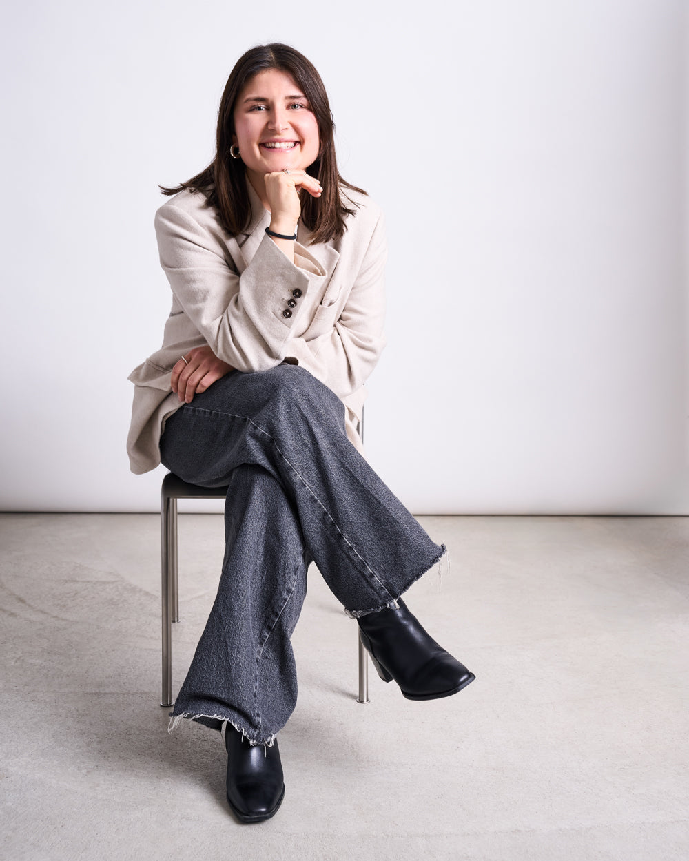Woman seated on a simple metal chair wearing a light beige blazer, dark grey frayed‑hem jeans and black square‑toe boots, one hand under her chin and the other resting on her leg.