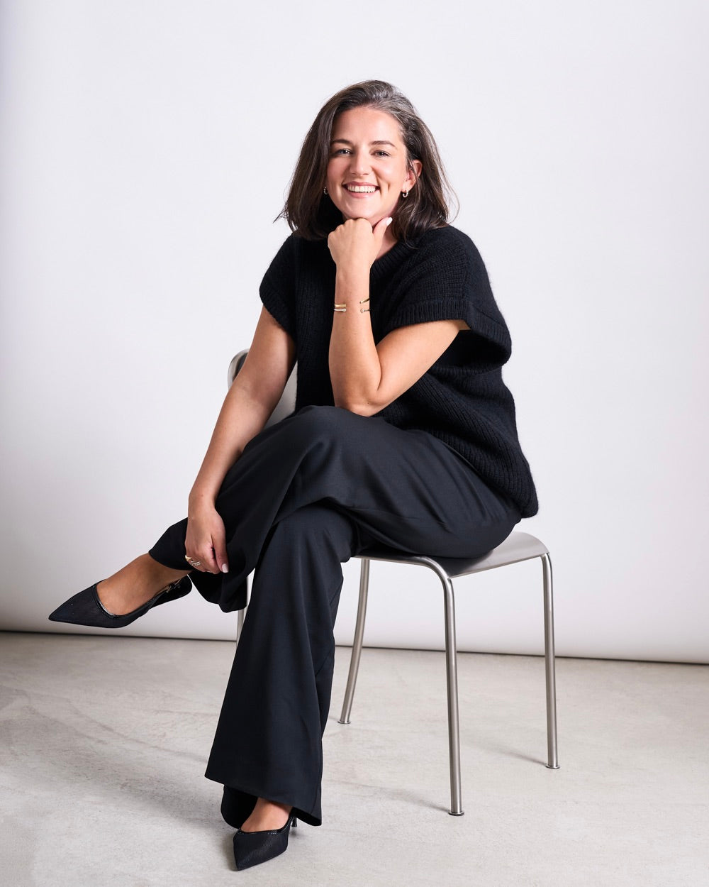 Woman seated on a metal chair wearing a black short‑sleeve knit top and wide‑leg black trousers, one leg crossed and hand resting under her chin.