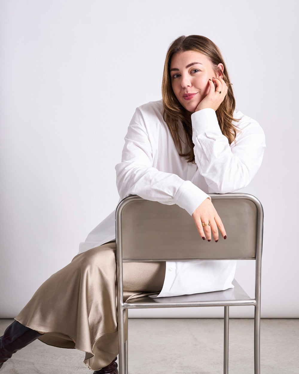 Woman seated behind a backward‑facing metal chair, wearing a long white shirt, a light beige satin skirt and dark boots, resting one hand on the backrest and the other supporting her head.