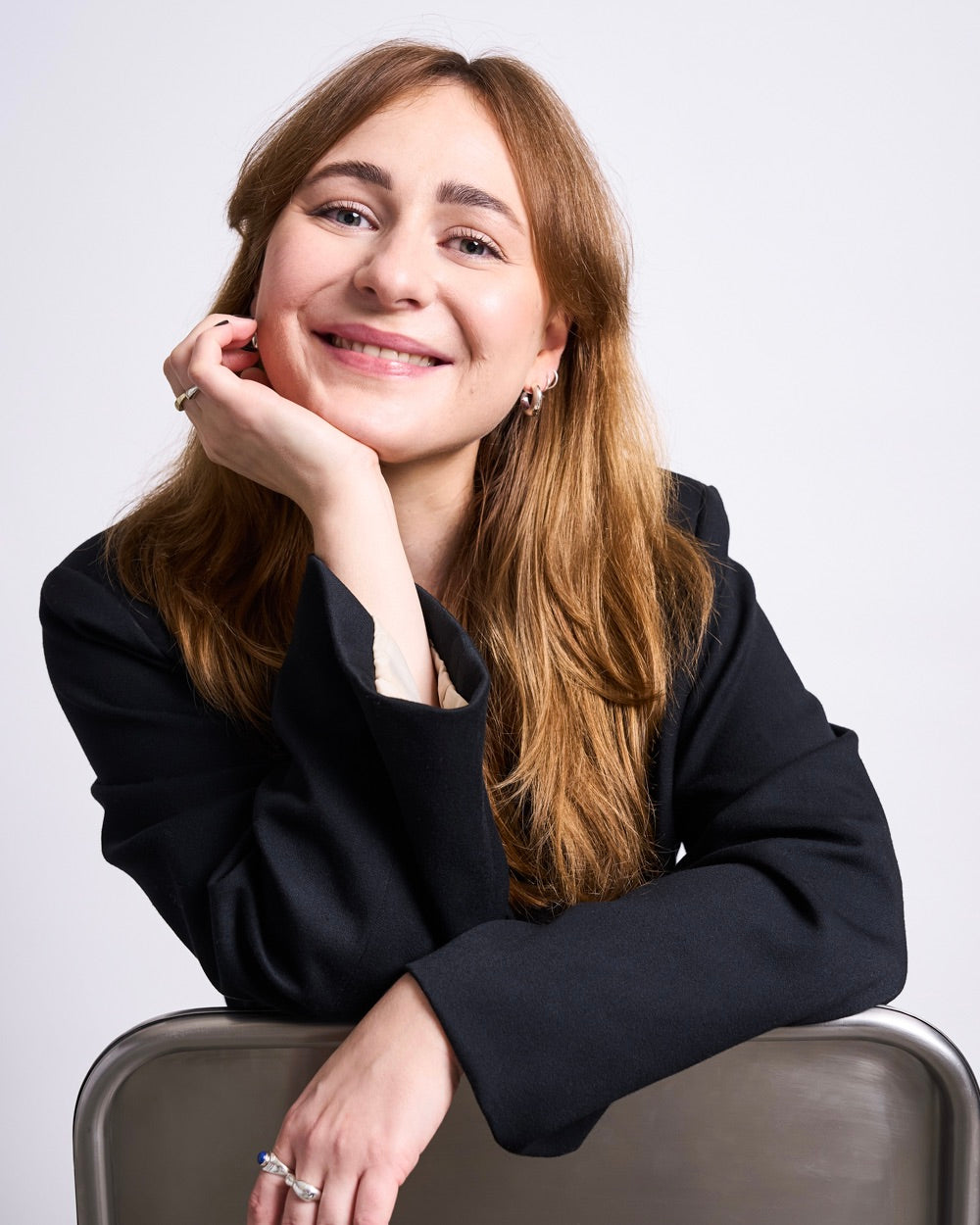 Person seated behind a metal chair wearing a dark blazer, head resting on one hand and smiling slightly, against a plain white background.