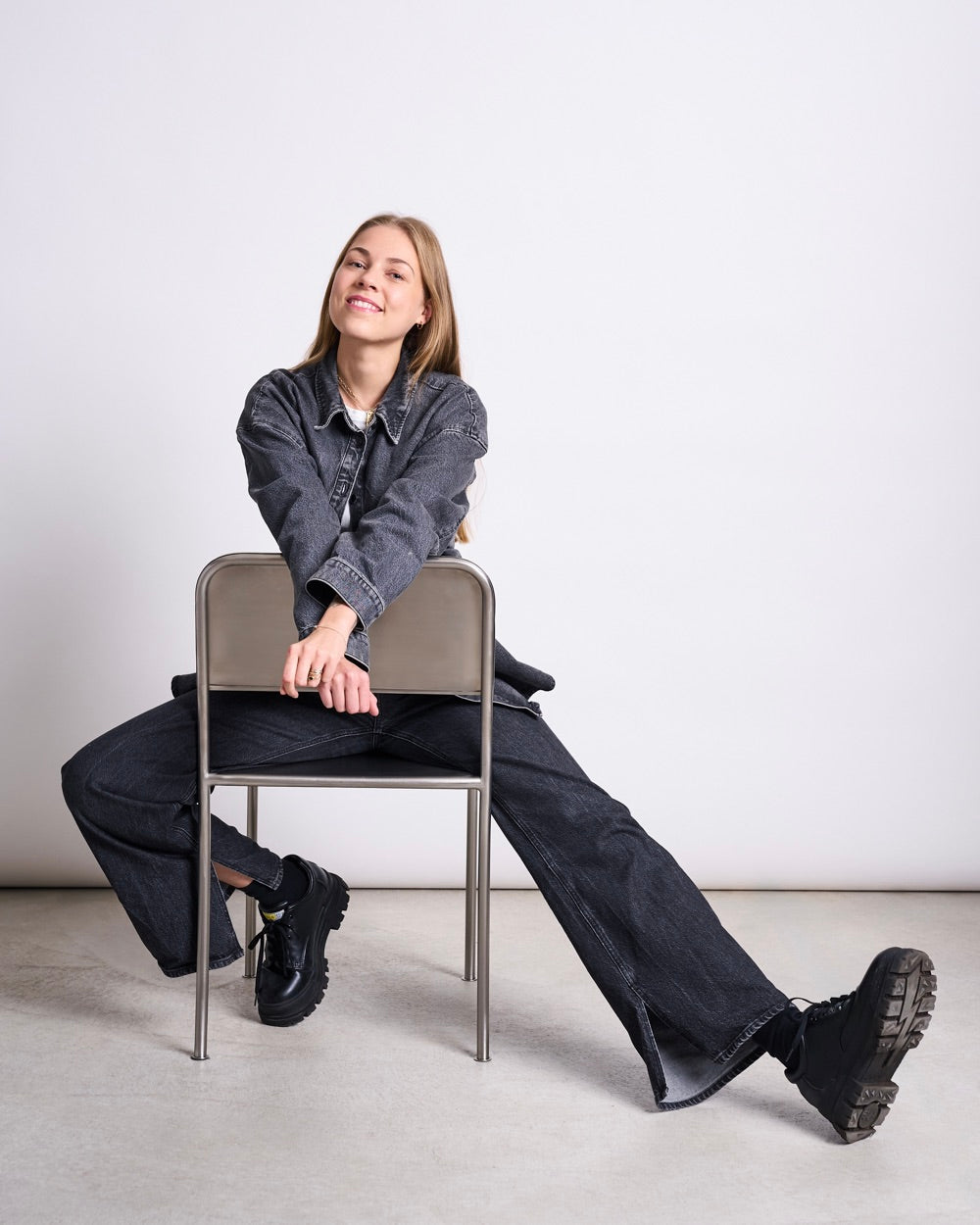 Women seated on a backward‑facing metal chair, wearing a dark denim jacket, matching dark denim trousers and black chunky shoes, arms resting on the backrest.