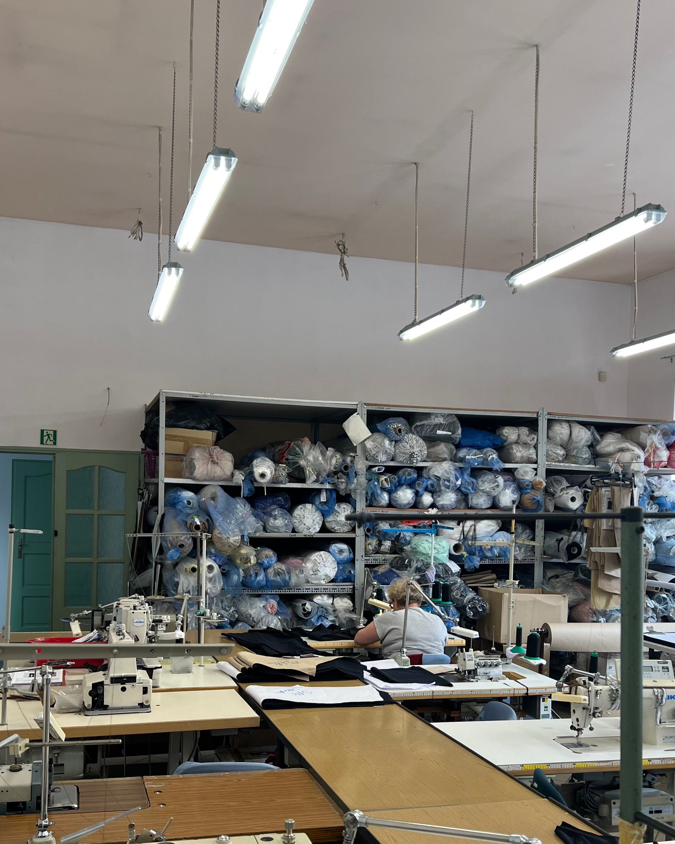 Interior of a textile workshop with industrial sewing machines on large tables, a person working with fabric in the foreground, and shelves filled with fabric rolls in the background, brightly lit by overhead lights.