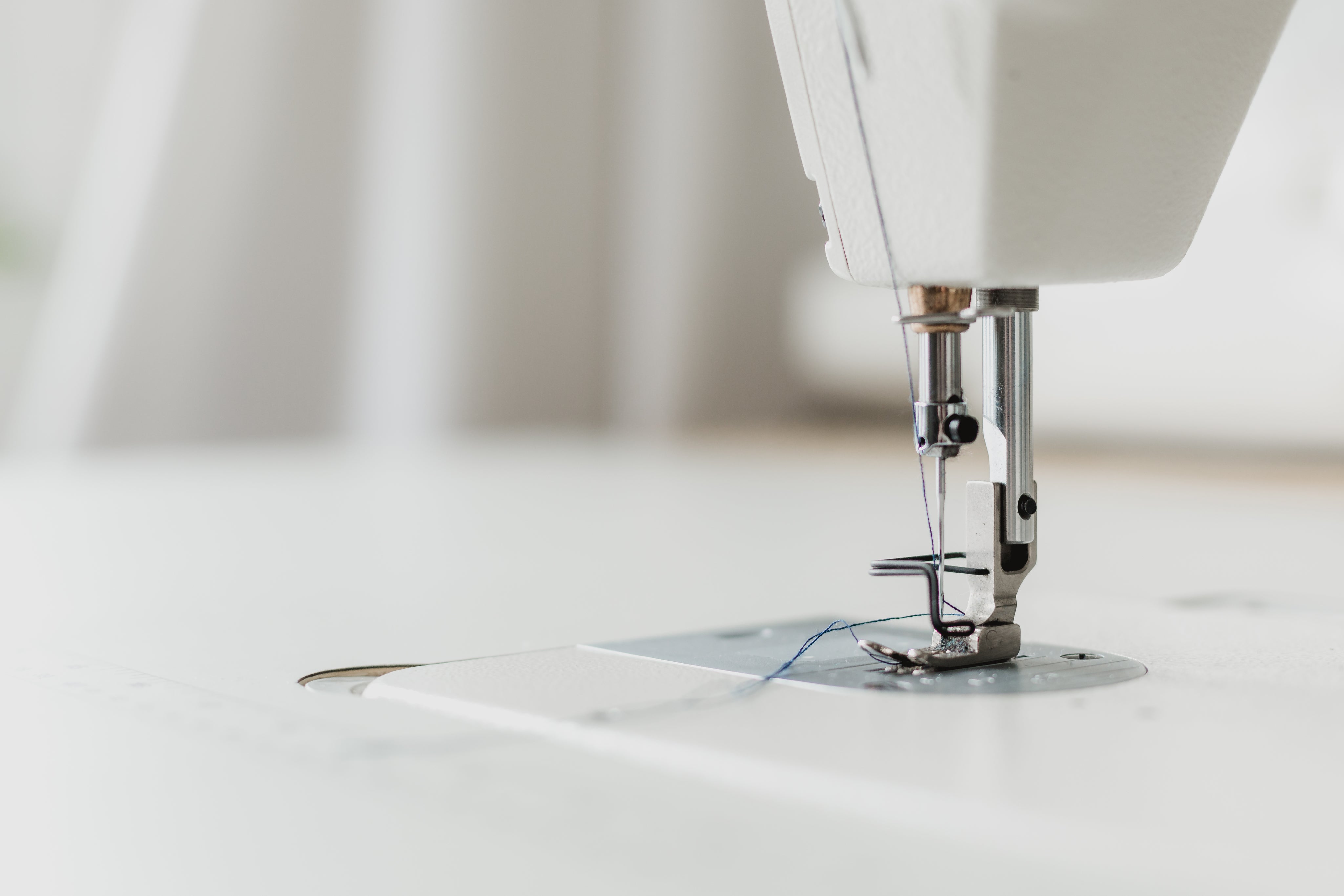 Close-up of a sewing machine needle and presser foot with a threaded needle, set against a blurred white background. The image focuses on the metal parts and details of the sewing machine.